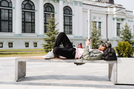 People, technology, travel and tourism - Afro man using smartphone on city street bench and texting on a sunny dayの写真素材