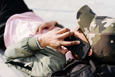 People, technology, travel and tourism - Afro man using smartphone on city street bench and texting on a sunny dayの写真素材