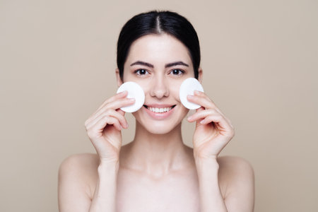Beautiful Caucasian woman gently cleaning face with makeup remover pad on beige backgroundの写真素材