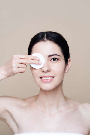 Beautiful Caucasian woman gently cleaning face with makeup remover pad on beige backgroundの写真素材