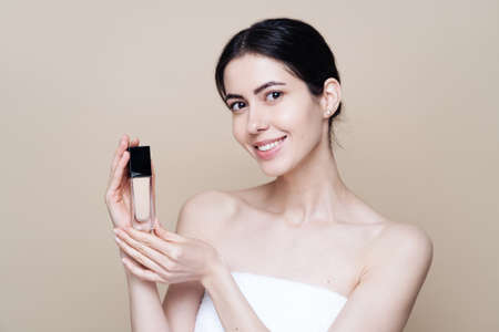 Woman holds Foundation. Photo of pretty woman with perfect skin on beige background. Beauty product...