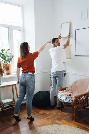 Excited african young husband and caucasian wife hang frame picture on wall happy settling together moving to new house, overjoyed biracial couple decorating own home relocating, ownership conceptの写真素材