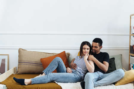 Portrait of happy young African Caucasian couple hug sitting on couch at home, smiling mixed race...