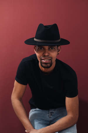 Portrait of handsome young African man smiling in black t-shirt and hat on burgundy backgroundの写真素材