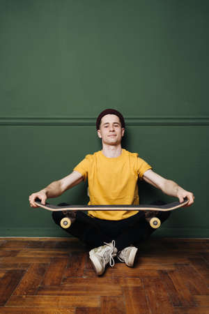 Portrait of young fashionable hipster man posing over green wall background. Caucasian male holding skateboard and smilingの写真素材