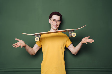 Portrait of young fashionable hipster man posing over green wall background. Caucasian male holding skateboard and smilingの写真素材