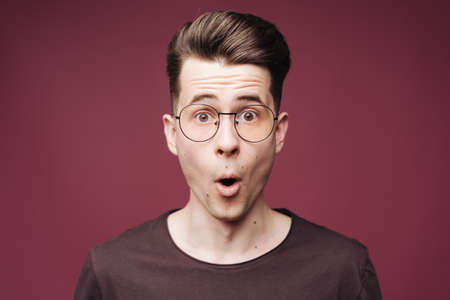 Surprised male, keeps mouth opened, being stupefied as advertises something, wears glasses, brown t shirt over crimson background. Omg conceptの写真素材