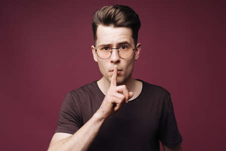 Portrait of emotional young guy holding index finger at his lips, saying shh, asking for silence or...