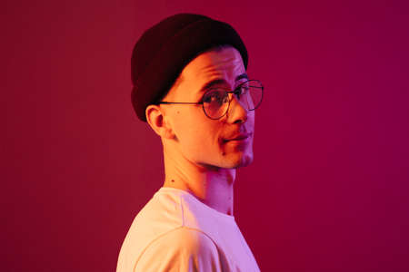 Shot of young handsome male with glasses trendy hairdo, wears casual white t shirt, has positive expression, poses in studio against pink background splashed by colorful lightsの写真素材