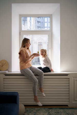 Daughter who loves her mother give her a potted plant present while sitting on the window at homeの写真素材