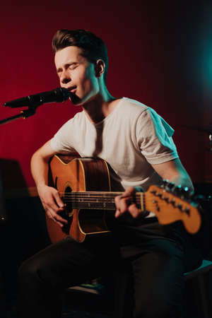 Talented handsome young guitarist man singing a song in studio recording on red background...