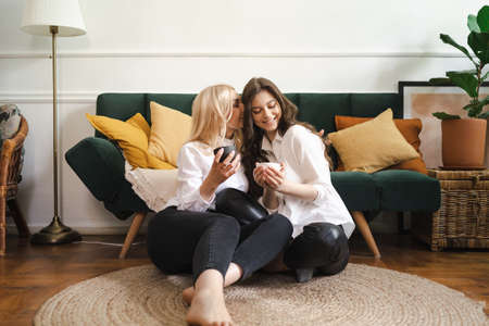 Beautiful young woman and adult girl are drinking tea, talking and smiling while sitting on floor at modern homeの写真素材