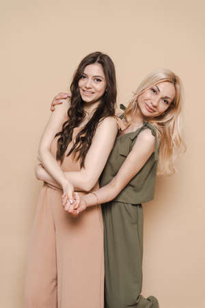 Blonde mother and brunette adult daughter wearing identical clothes, different colors, posing over beige backgroundの写真素材