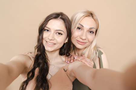 Portrait of beautiful blonde mother and her daughter making a selfie using smart phone and smiling, on beige background.の写真素材