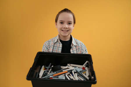 Smiling School boy holding a box with plastic robotic peaces on yellow wall background. Ready to...