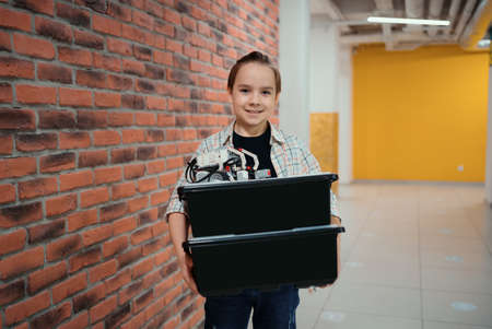 Smiling School boy walking while holding boxes with plastic robotic peaces on brick wall background. Ready to create. Adventure time.の写真素材