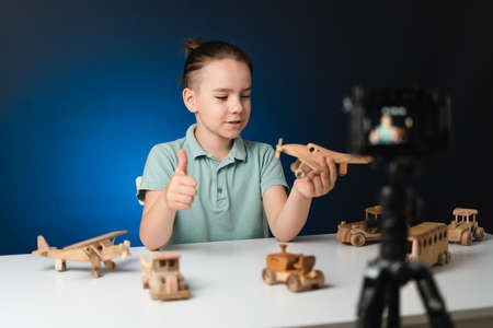 Kid blogger filming video on camera indoors with blue light in background. Online reviewing. Promoting eco wooden toys. Kid recording his gameplay for vlog.の写真素材