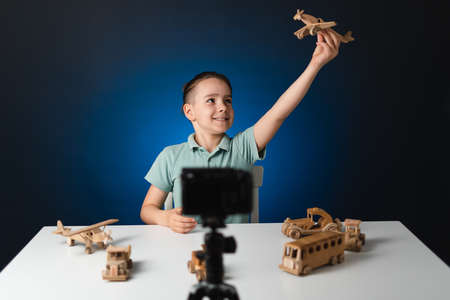 Kid blogger filming video on camera indoors with blue light in background. Online reviewing. Promoting eco wooden toys. Kid recording his gameplay for vlog.の写真素材