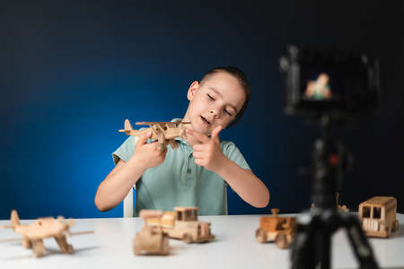 Kid blogger filming video on camera indoors with blue light in background. Online reviewing. Promoting eco wooden toys. Kid recording his gameplay for vlog.の写真素材
