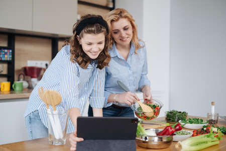 Mom and daughter cooking together in the kitchen using a receipt on tablet. They use vegetables for cooking in a good mood, happy to be togetherの写真素材