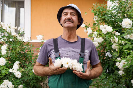 Smiling man portrait with a white rose on his hat with bushes of roses in background