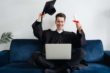 Education, graduation and people concept - happy male student with diploma and laptop at home showing his emotionsの写真素材