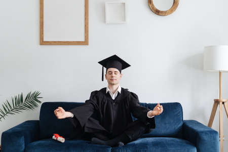 Education, graduation and people concept - happy male student sitting in yoga pose on blue couch at home showing his emotions. Meditationの写真素材