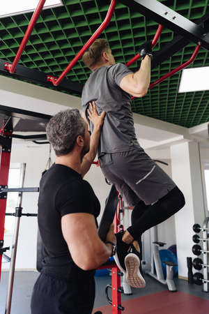 A personal trainer motivates the client to do liftings on horizontal bar in the gym. Personal workout hour with personal coachの写真素材