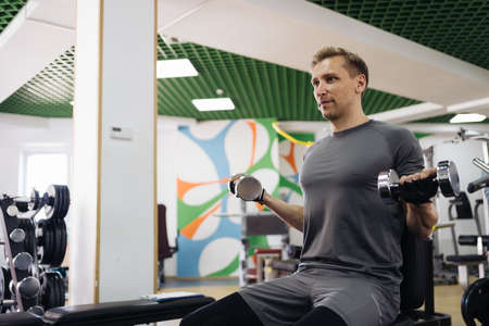 Attractive Caucasian blonde man lift heavy dumbbells in the gym. Personal workout hour. Daily sport...