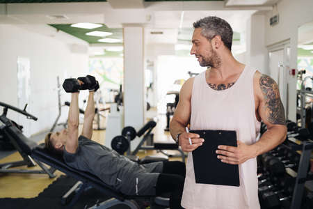 Muscular handsome trainer writing a fitness plan on clipboard for working out near his trainee in...