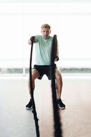 Caucasian blonde man training with rope in functional training fitness gym with big windows and a...