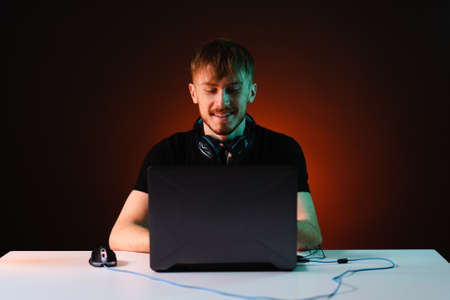 Portrait of Software Developer. Hacker. Gamer Wearing Headset Sitting at His Desk and Working. Playing on Laptop. In the Background Dark High Tech Environment with red neon lights.の写真素材