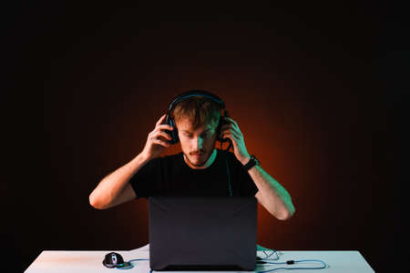 Portrait of Software Developer. Hacker. Gamer Wearing Headset Sitting at His Desk and Working. Playing on Laptop. In the Background Dark High Tech Environment with red neon lights.の写真素材
