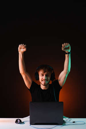 Joyful young gamer rejoices in winning a video game with a smile on his face and raised hands, wears casual clothes and a headset. Streamer winner plays at home on the computer on dark background.の写真素材