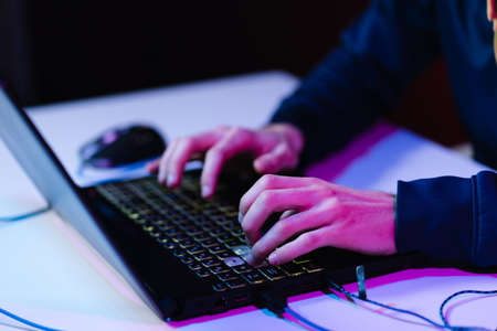 Hacker with a laptop sits at the table, dark background and neon light behind, head covered with a hood.の写真素材