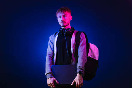 Neon portrait of a bearded man wearing headphones on the neck, parka, backpack, mate sunglasses holding a gamer laptop in hands. Hacker portraitの写真素材