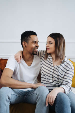 Portrait of happy young African Caucasian couple hug sitting on couch at home, smiling mixed race husband and wife spend time together, embrace relaxing on sofa. Healthy relationships conceptの写真素材