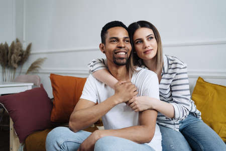 Portrait of happy young African Caucasian couple hug sitting on couch at home, smiling mixed race...