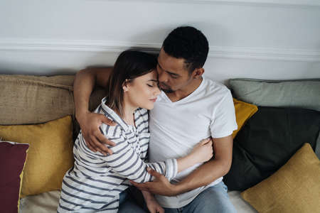 Portrait of happy young African Caucasian couple hug sitting on couch at home, smiling mixed race...