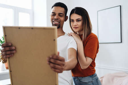 Excited african young husband and caucasian wife hang frame picture on wall happy settling together moving to new house, overjoyed biracial couple decorating own home relocating, ownership conceptの写真素材