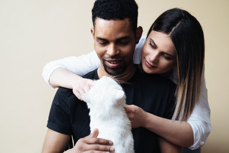 Lovely mixed-races young happy joyful couple male and female smiling in good mood resting with pet animal holding cat in hands, pet lover concept on beige backgroundの写真素材