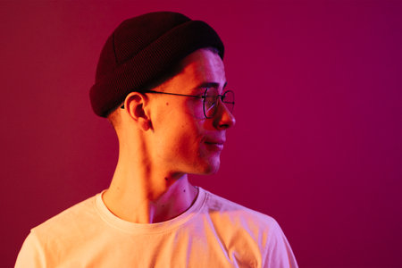 Shot of young handsome male with glasses trendy hairdo, wears casual white t shirt, has positive expression, poses in studio against pink background splashed by colorful lightsの写真素材