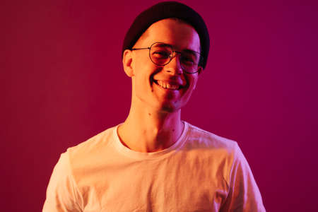 Shot of young handsome male with glasses trendy hairdo, wears casual white t shirt, has positive expression, poses in studio against pink background splashed by colorful lightsの写真素材
