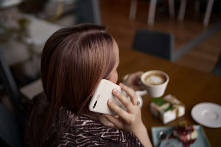 Close-up portrait of smiling woman drinking coffee alone while talking on smartphone. Lovable lady standing near brick wall in a cafeの写真素材