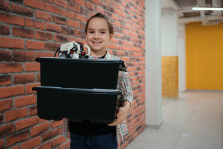 Smiling School boy walking while holding boxes with plastic robotic peaces on brick wall background....