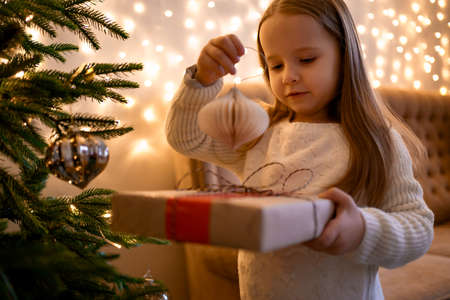 Wondering little girl looking enthusiastic at a present standing near decorated fire tree, Led lights and sofa in the backgroundの写真素材
