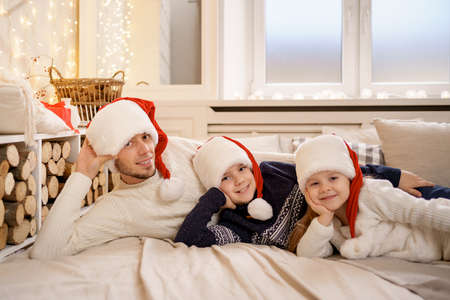 Merry Christmas and Happy New Year. Happy family are waiting Xmas Eve in Santa Claus hats while lying on bed near the window at home.の写真素材