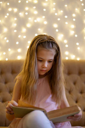 Girl with long wavy hair reading an old book with fairy tales while sitting on sofa with lights in backgroundの写真素材