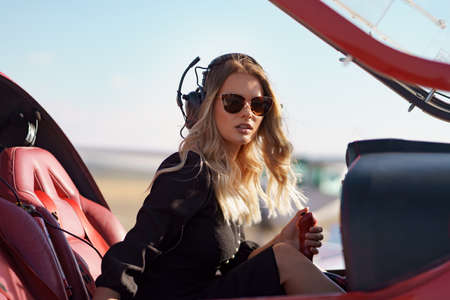 Young fashionable woman pilot in headset ready to fly in small red airplane. Beautiful life, aristocratic lady in black dress in blonde wavy hair.の写真素材