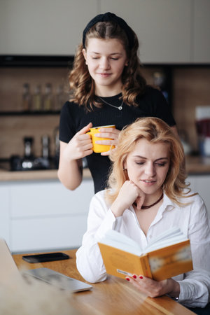 Mother discussing with her daughter teenager a book while drinking tea in living room at homeの写真素材